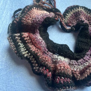 May include: A handmade crochet scarf in shades of black, pink, purple, and white. The scarf has a textured, wavy design and is made of soft yarn. The ends of the scarf have long, dangling yarn strands.