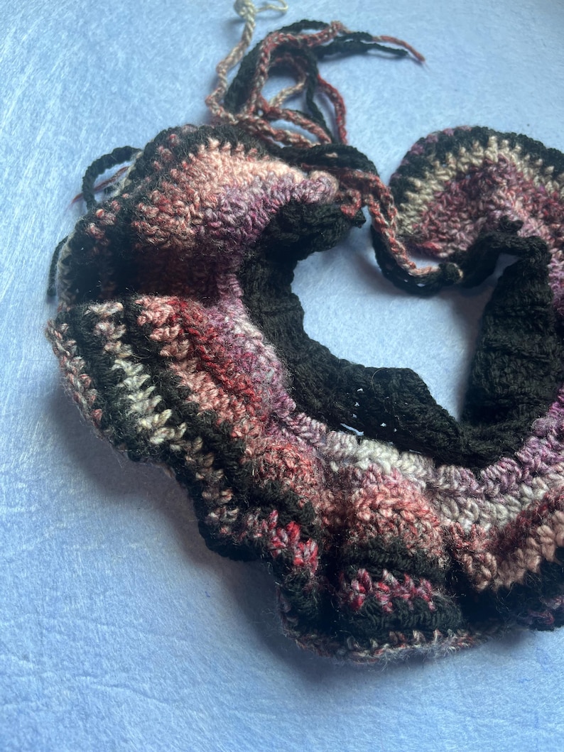 May include: A handmade crochet scarf in shades of black, pink, purple, and white. The scarf has a textured, wavy design and is made of soft yarn. The ends of the scarf have long, dangling yarn strands.
