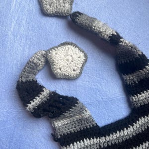 May include: Crocheted cat tail and two star-shaped pieces in shades of gray and black. The tail is striped with black, gray, and white. The star-shaped pieces are gray and white. The items are handmade and likely part of a costume or toy.