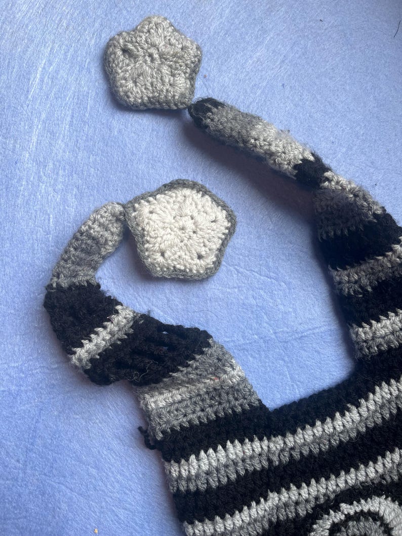 May include: Crocheted cat tail and two star-shaped pieces in shades of gray and black. The tail is striped with black, gray, and white. The star-shaped pieces are gray and white. The items are handmade and likely part of a costume or toy.