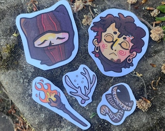May include: A collection of five fantasy-themed stickers. The stickers feature illustrations of a hooded figure, a person with closed eyes, a torch with flames, a coral-like shape, and a hat with text.