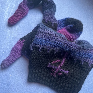 May include: A handmade crochet hat in shades of black, purple, and blue. The hat features a long, curved extension and a stitched symbol on the front. The hat is made of soft yarn and is suitable for costume or everyday wear.