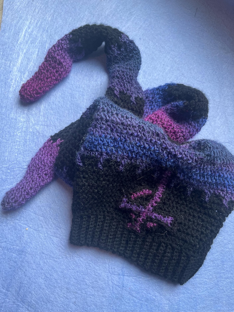 May include: A handmade crochet hat in shades of black, purple, and blue. The hat features a long, curved extension and a stitched symbol on the front. The hat is made of soft yarn and is suitable for costume or everyday wear.
