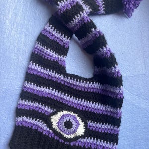 May include: A handmade crochet hat in black, purple, and white stripes, featuring a large eye design. The hat has a long, striped strap with purple pom-poms at the ends. The hat is designed to be worn on the head.