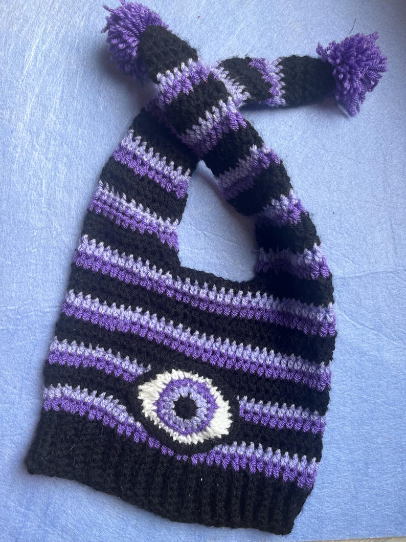 May include: A handmade crochet hat in black, purple, and white stripes, featuring a large eye design. The hat has a long, striped strap with purple pom-poms at the ends. The hat is designed to be worn on the head.