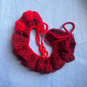 May include: A handmade, red crocheted collar and matching cuff. The collar has a ruffled edge and a small flower detail. The cuff is a small, rounded shape with a drawstring. Both items are made of a textured yarn.