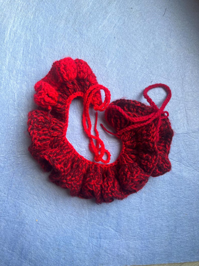 May include: A handmade, red crocheted collar and matching cuff. The collar has a ruffled edge and a small flower detail. The cuff is a small, rounded shape with a drawstring. Both items are made of a textured yarn.