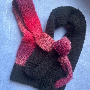 May include: A handmade crochet scarf in black, with a gradient of pink and red. The scarf features a loop and a pom-pom detail. The scarf is made of a soft, textured yarn, perfect for cold weather. The scarf is a cozy and stylish accessory.