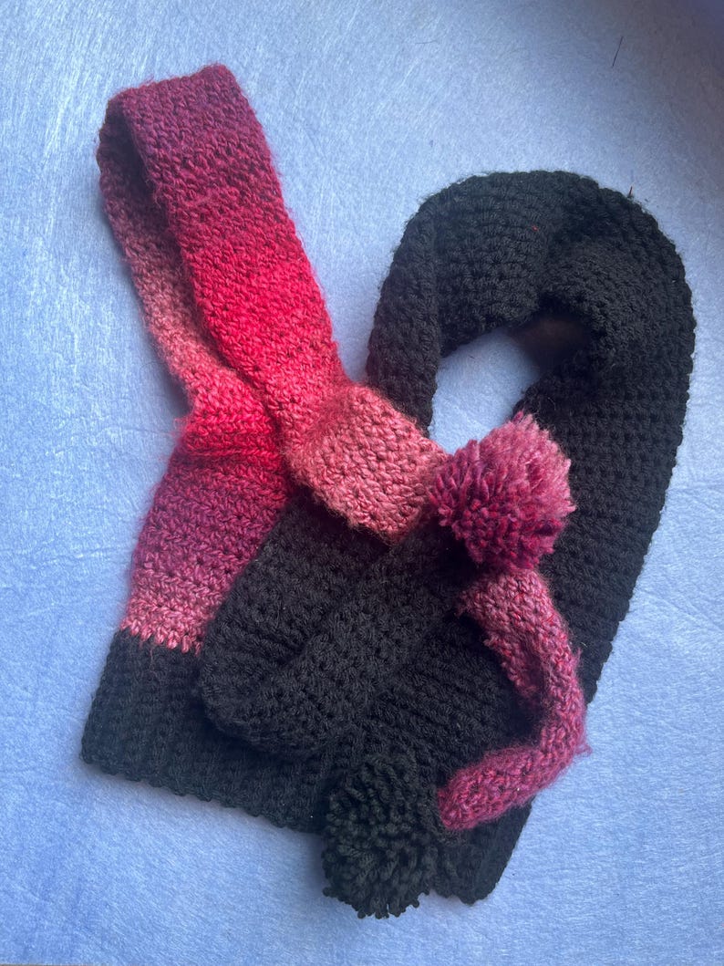 May include: A handmade crochet scarf in black, with a gradient of pink and red. The scarf features a loop and a pom-pom detail. The scarf is made of a soft, textured yarn, perfect for cold weather. The scarf is a cozy and stylish accessory.