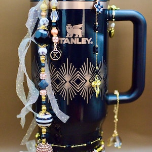 Puede incluir: Un termo Stanley negro con diseños geométricos dorados y el nombre de la marca. El termo está adornado con abalorios de cuentas en varios colores, incluyendo negro, dorado y cristales transparentes. Una pieza decorativa con la palabra "DECO" envuelve la base.