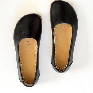 May include: A pair of black leather flats with a rounded toe and a tan insole. The shoes have a stitched design around the sole and a small copper-coloured accent on the insole. The shoes are displayed on a white background.