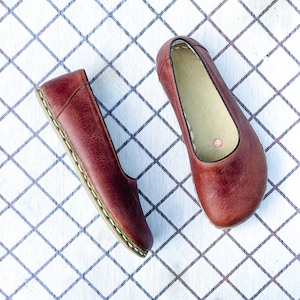 May include: A pair of burgundy leather ballet flats. The shoes have a rounded toe and a stitched sole. The interior is a light beige color. The shoes are displayed on a white surface with a blue grid pattern.