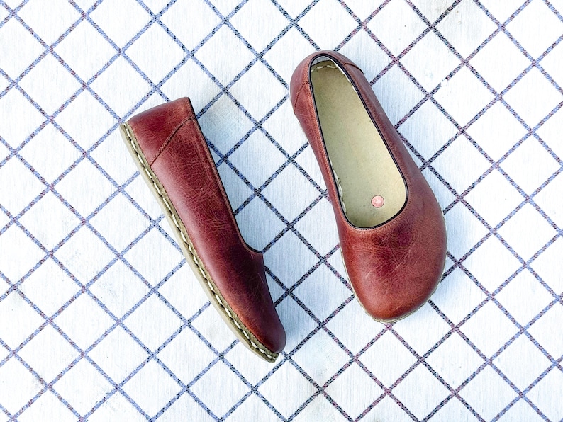 May include: A pair of burgundy leather ballet flats. The shoes have a rounded toe and a stitched sole. The interior is a light beige color. The shoes are displayed on a white surface with a blue grid pattern.