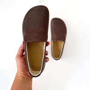 Pu&ograve; includere: Un paio di scarpe slip-on in pelle marrone. Le scarpe hanno la punta arrotondata e una superficie strutturata. L'interno &egrave; di colore beige chiaro. Le scarpe sono esposte su sfondo bianco.