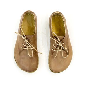 Può includere: Un paio di scarpe in pelle marrone con lacci beige, viste dall'alto. Le scarpe hanno un classico stile Oxford con punta arrotondata e suola cucita. L'interno è di colore marrone chiaro.