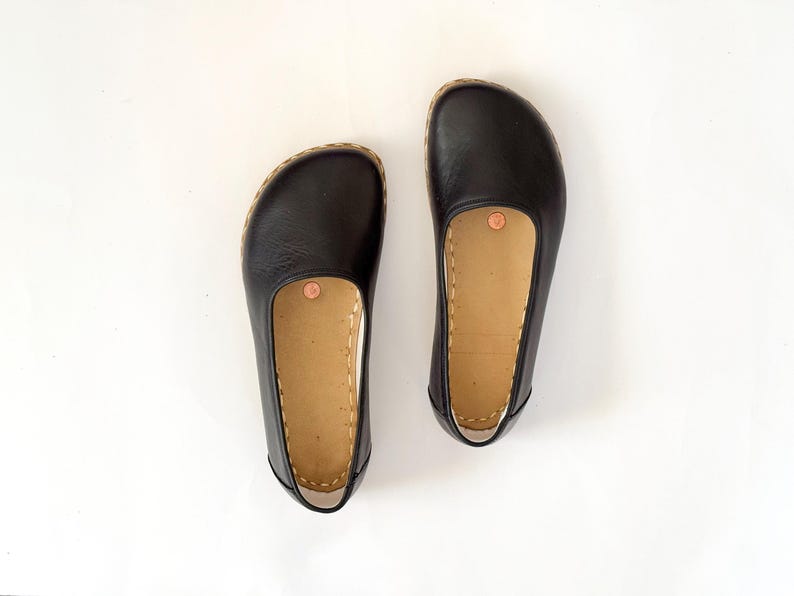 May include: A pair of black leather flats with a rounded toe and a tan insole. The shoes have a stitched sole and a copper-coloured rivet on the insole. The shoes are displayed on a white background.