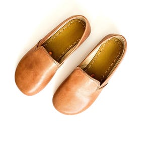 Pode incluir: Um par de sapatos slip-on de couro castanho. Os sapatos t&ecirc;m bico arredondado e perfil baixo. O interior &eacute; castanho mais claro, com costuras vis&iacute;veis nas bordas. Sapatos confort&aacute;veis para uso di&aacute;rio.
