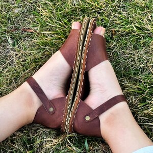 Pu&ograve; includere: Sandali in pelle marrone con anello per le dita e cinturino alla caviglia. I sandali hanno una suola cucita e sono mostrati su una superficie erbosa. I sandali sono di un caldo colore marrone.