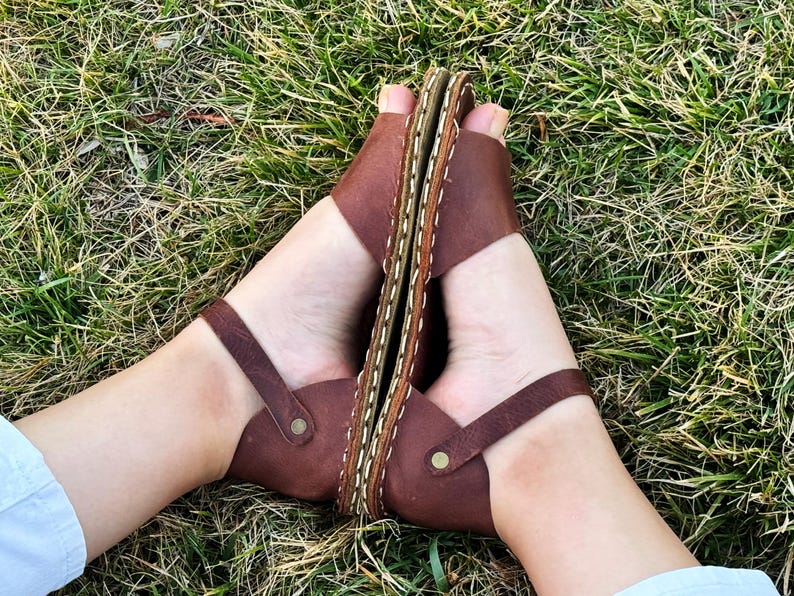 Pu&ograve; includere: Sandali in pelle marrone con anello per le dita e cinturino alla caviglia. I sandali hanno una suola cucita e sono mostrati su una superficie erbosa. I sandali sono di un caldo colore marrone.