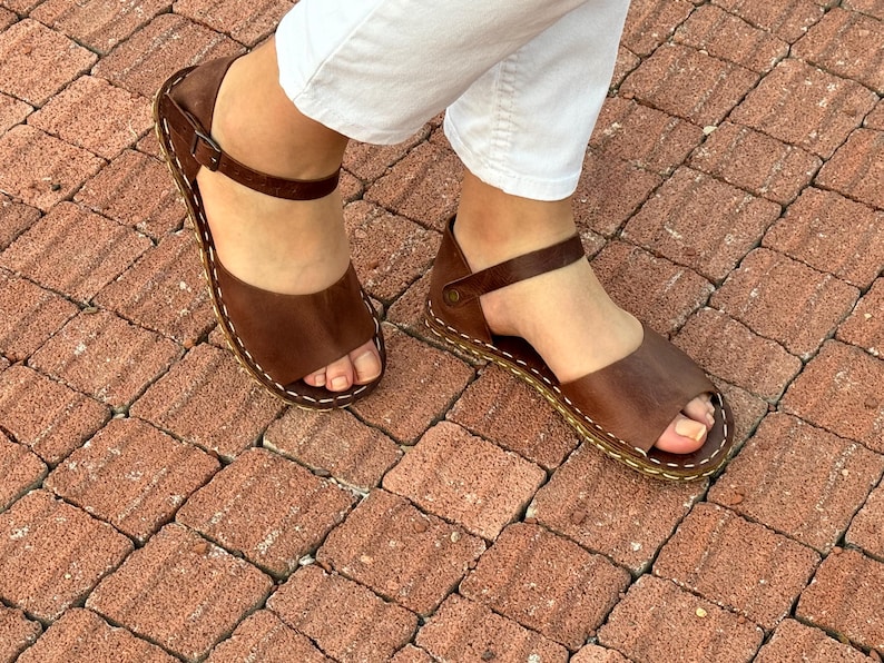 Pu&ograve; includere: Sandali in pelle marrone con design a punta aperta e cinturino alla caviglia. I sandali hanno cuciture bianche sui bordi e sono indossati con pantaloni bianchi. Su una superficie di mattoni rossi.