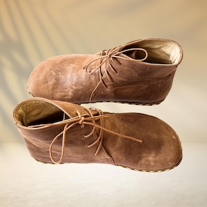 May include: A pair of brown leather ankle boots with laces. The boots have a worn, textured appearance and are set against a neutral background with a subtle shadow.