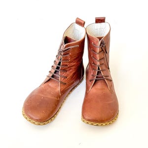 May include: A pair of brown leather ankle boots with a lace-up design. The boots have a rounded toe and a pull tab at the top. The soles are a lighter brown color.