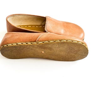 Pode incluir: Um par de sapatos slip-on de couro castanho claro. Os sapatos t&ecirc;m bico arredondado e sola cosida. As solas s&atilde;o de cor castanho mais escuro. Os sapatos s&atilde;o mostrados em fundo branco.