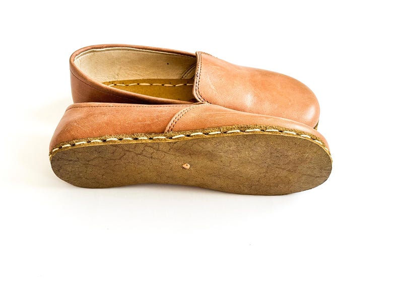 Pode incluir: Um par de sapatos slip-on de couro castanho claro. Os sapatos t&ecirc;m bico arredondado e sola cosida. As solas s&atilde;o de cor castanho mais escuro. Os sapatos s&atilde;o mostrados em fundo branco.
