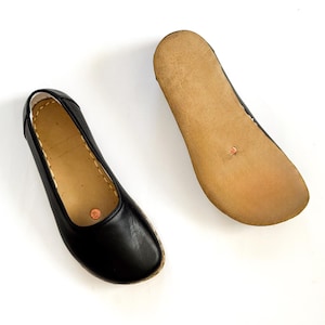 May include: A pair of black leather ballet flats. One shoe is right-side up, showing the top and sides, while the other is upside down, revealing the light brown sole. The shoes have a simple, classic design.