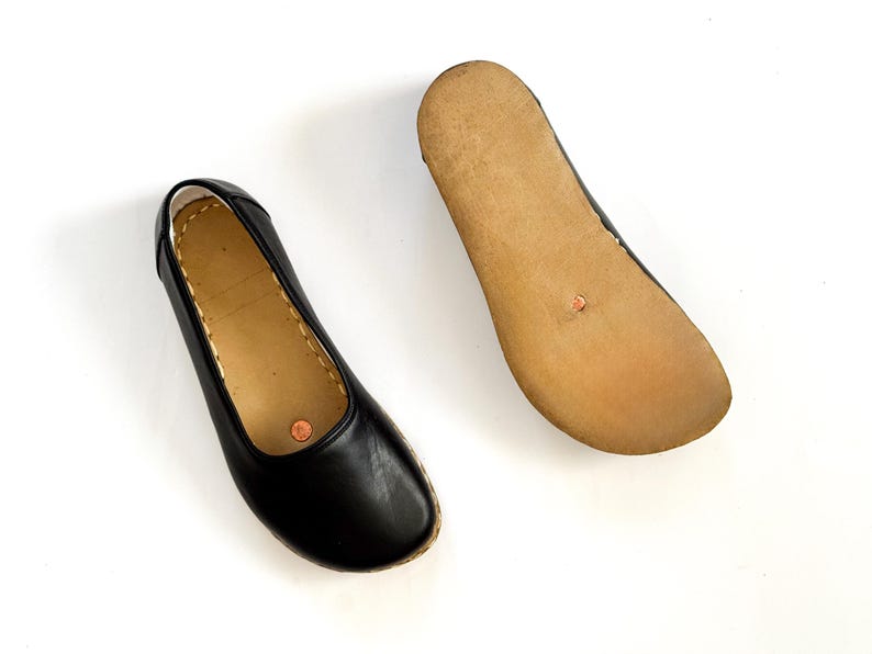 May include: A pair of black leather ballet flats. One shoe is right-side up, showing the top and sides, while the other is upside down, revealing the light brown sole. The shoes have a simple, classic design.