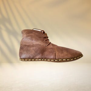 Puede incluir: Una bota de cuero marr&oacute;n con dise&ntilde;o de cordones. La bota tiene una superficie texturizada y una suela cosida. El fondo es beige neutro con la sombra de una hoja de palma.