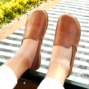 Pode incluir: Um par de sapatos slip-on de couro castanho. Os sapatos t&ecirc;m bico arredondado e perfil baixo. A imagem mostra os sapatos de uma perspectiva de cima para baixo, com os p&eacute;s do usu&aacute;rio vis&iacute;veis. Os sapatos s&atilde;o de uma cor castanha quente.