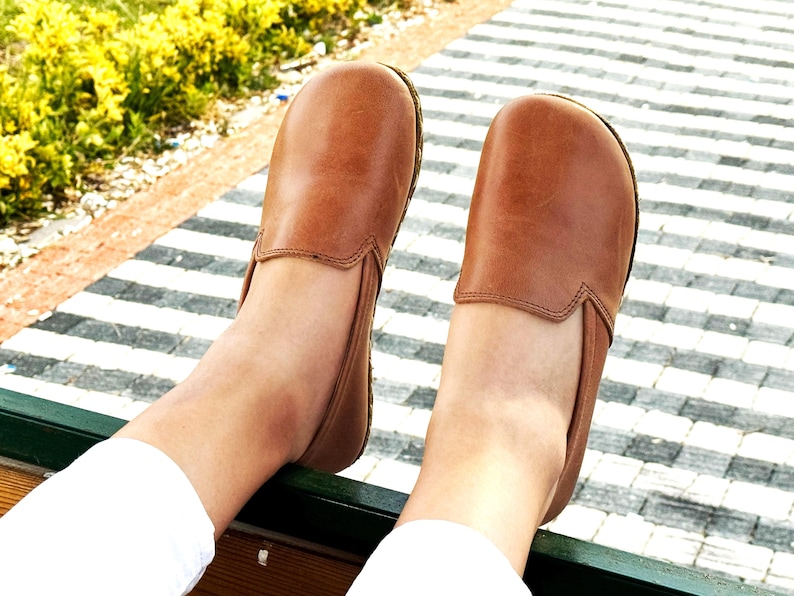 Pode incluir: Um par de sapatos slip-on de couro castanho. Os sapatos t&ecirc;m bico arredondado e perfil baixo. A imagem mostra os sapatos de uma perspectiva de cima para baixo, com os p&eacute;s do usu&aacute;rio vis&iacute;veis. Os sapatos s&atilde;o de uma cor castanha quente.