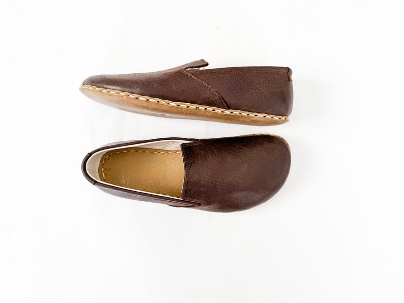 Pu&ograve; includere: Due scarpe marroni in pelle senza lacci. Le scarpe hanno la punta arrotondata e la suola cucita. L'interno della scarpa &egrave; di colore beige chiaro. Le scarpe sono esposte su sfondo bianco.