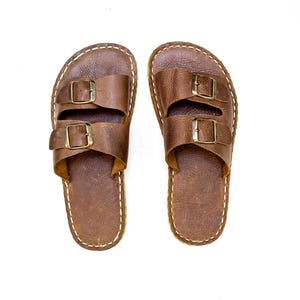 Op de afbeelding: Een paar bruine leren sandalen met twee gespen op elke band.