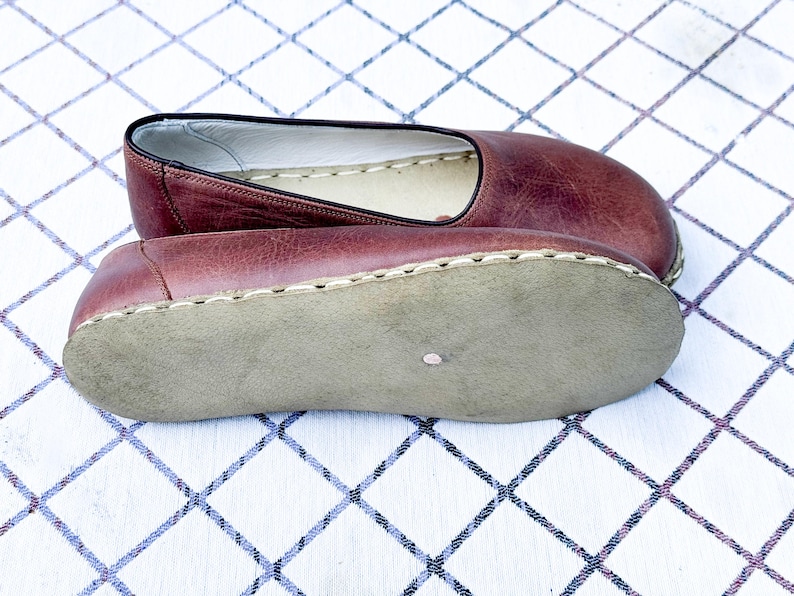 May include: A pair of burgundy leather slip-on shoes. The shoes have a rounded toe and a stitched sole. The soles are a light tan color. The shoes are displayed on a white and blue checkered surface.