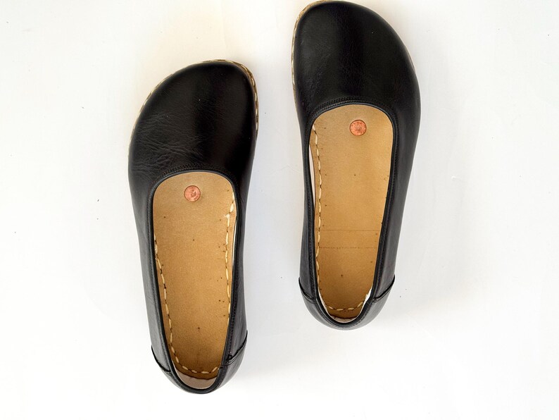 May include: A pair of black leather flats with a rounded toe and a tan insole. The shoes have a stitched design around the sole and a small copper-coloured accent on the insole. The shoes are displayed on a white background.