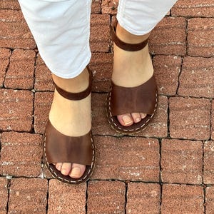 Pu&ograve; includere: Sandali in pelle marrone con cinturini alla caviglia. I sandali hanno un design a punta aperta e sono cuciti con filo bianco. L'immagine mostra i sandali da una prospettiva dall'alto, indossati con pantaloni bianchi.