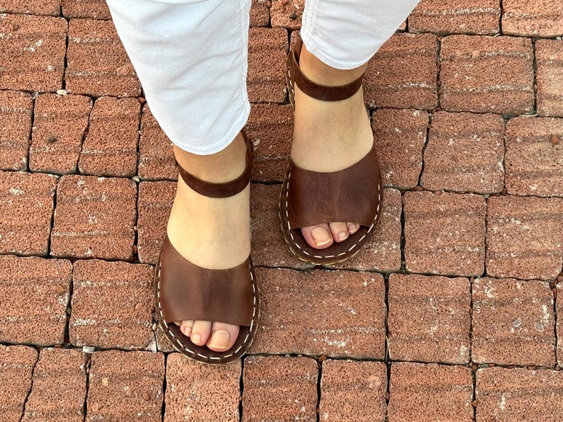 Pu&ograve; includere: Sandali in pelle marrone con cinturini alla caviglia. I sandali hanno un design a punta aperta e sono cuciti con filo bianco. L'immagine mostra i sandali da una prospettiva dall'alto, indossati con pantaloni bianchi.