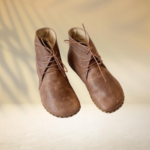 Puede incluir: Un par de botines de cuero marr&oacute;n con cordones. Los botines tienen una punta redondeada y un dise&ntilde;o texturizado en el lateral. Las suelas son de color marr&oacute;n claro. El fondo es un degradado de beige y bronceado.