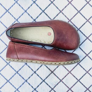 May include: A pair of burgundy leather flats with a rounded toe and a stitched sole. The shoes are displayed on a white surface with a blue and white diamond pattern.