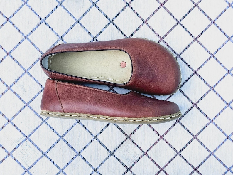 May include: A pair of burgundy leather flats with a rounded toe and a stitched sole. The shoes are displayed on a white surface with a blue and white diamond pattern.