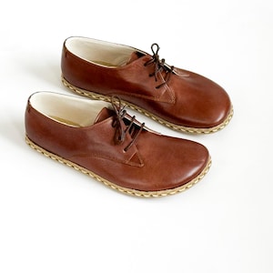 Peut inclure: Une paire de chaussures en cuir marron avec des lacets, sur fond blanc. Les chaussures ont un bout arrondi et une semelle cousue de couleur claire. La doublure int&eacute;rieure est blanche.