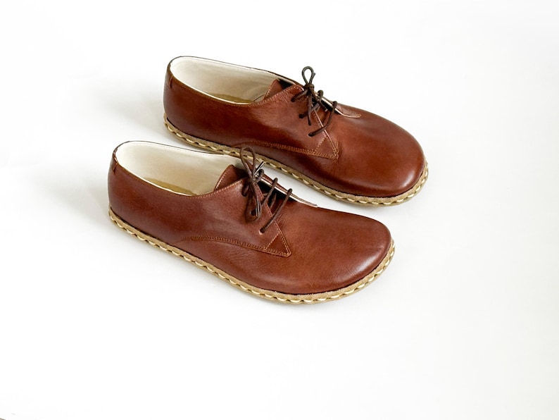 Peut inclure: Une paire de chaussures en cuir marron avec des lacets, sur fond blanc. Les chaussures ont un bout arrondi et une semelle cousue de couleur claire. La doublure int&eacute;rieure est blanche.