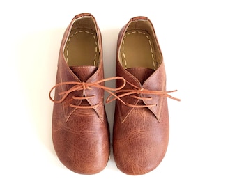 Peut inclure: Une paire de chaussures en cuir marron avec des lacets, sur fond blanc. Les chaussures ont un design classique avec un bout arrondi et des coutures visibles. Le cuir a un aspect texturé.