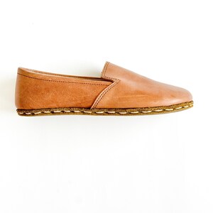Pode incluir: Um sapato slip-on de couro castanho. O sapato apresenta uma biqueira arredondada, uma sola cosida e um perfil baixo. O couro tem um aspeto natural, ligeiramente envelhecido. O sapato foi concebido para conforto e uso casual.