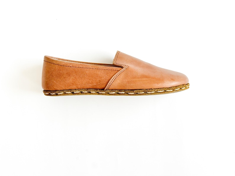 Pode incluir: Um sapato slip-on de couro castanho. O sapato apresenta uma biqueira arredondada, uma sola cosida e um perfil baixo. O couro tem um aspeto natural, ligeiramente envelhecido. O sapato foi concebido para conforto e uso casual.