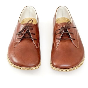Peut inclure: Une paire de chaussures en cuir marron avec des lacets, sur fond blanc. Les chaussures ont un bout arrondi et une semelle cousue. Le cuir a un aspect naturel et textur&eacute;.
