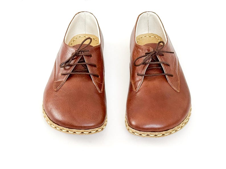 Peut inclure: Une paire de chaussures en cuir marron avec des lacets, sur fond blanc. Les chaussures ont un bout arrondi et une semelle cousue. Le cuir a un aspect naturel et textur&eacute;.