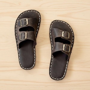 Pode incluir: Um par de sandálias de couro castanho escuro com duas tiras e fivelas prateadas. As sandálias têm costuras brancas nas bordas e estão sobre um fundo claro de grão de madeira. Calçado.